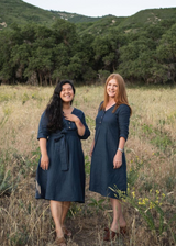 Two women in a field wearing the navy blue Faye linen dress with V-neck buttons and optional waist tie.