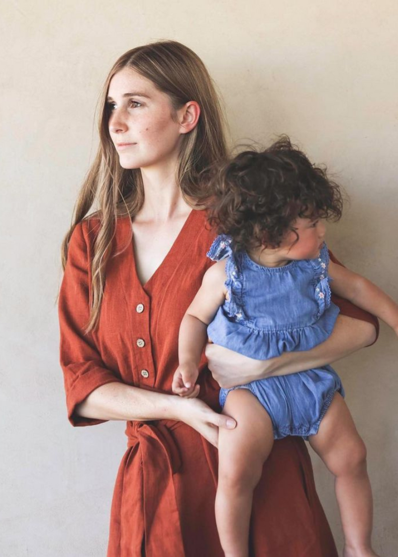 Woman in a rust linen Faye Dress with front buttons and waist tie, holding a baby.