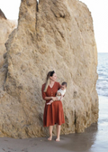 Woman in a rust linen Faye Dress with front buttons and V-neck, holding a baby on a beach.