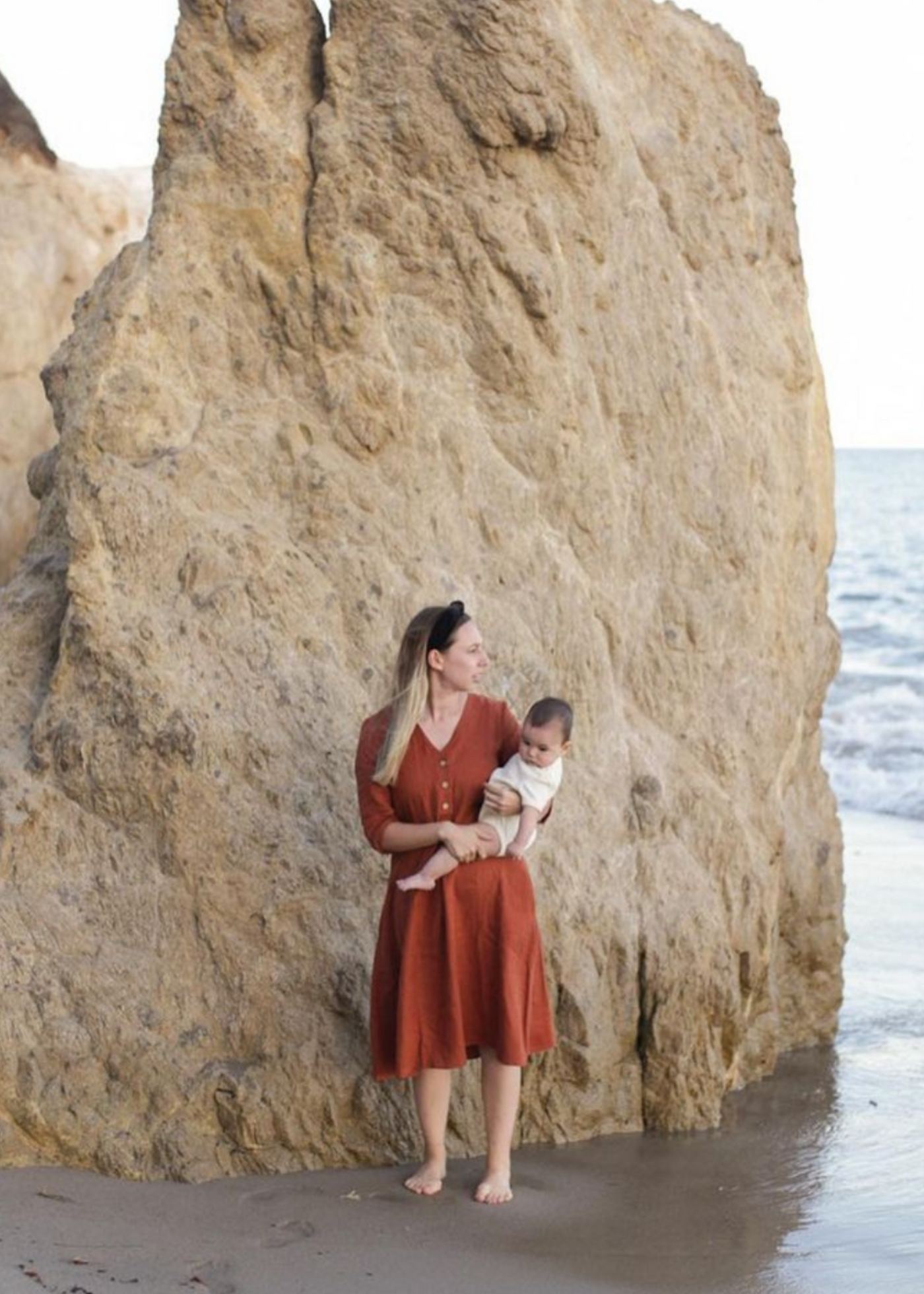 Woman in a rust linen Faye Dress with front buttons and V-neck, holding a baby on a beach.