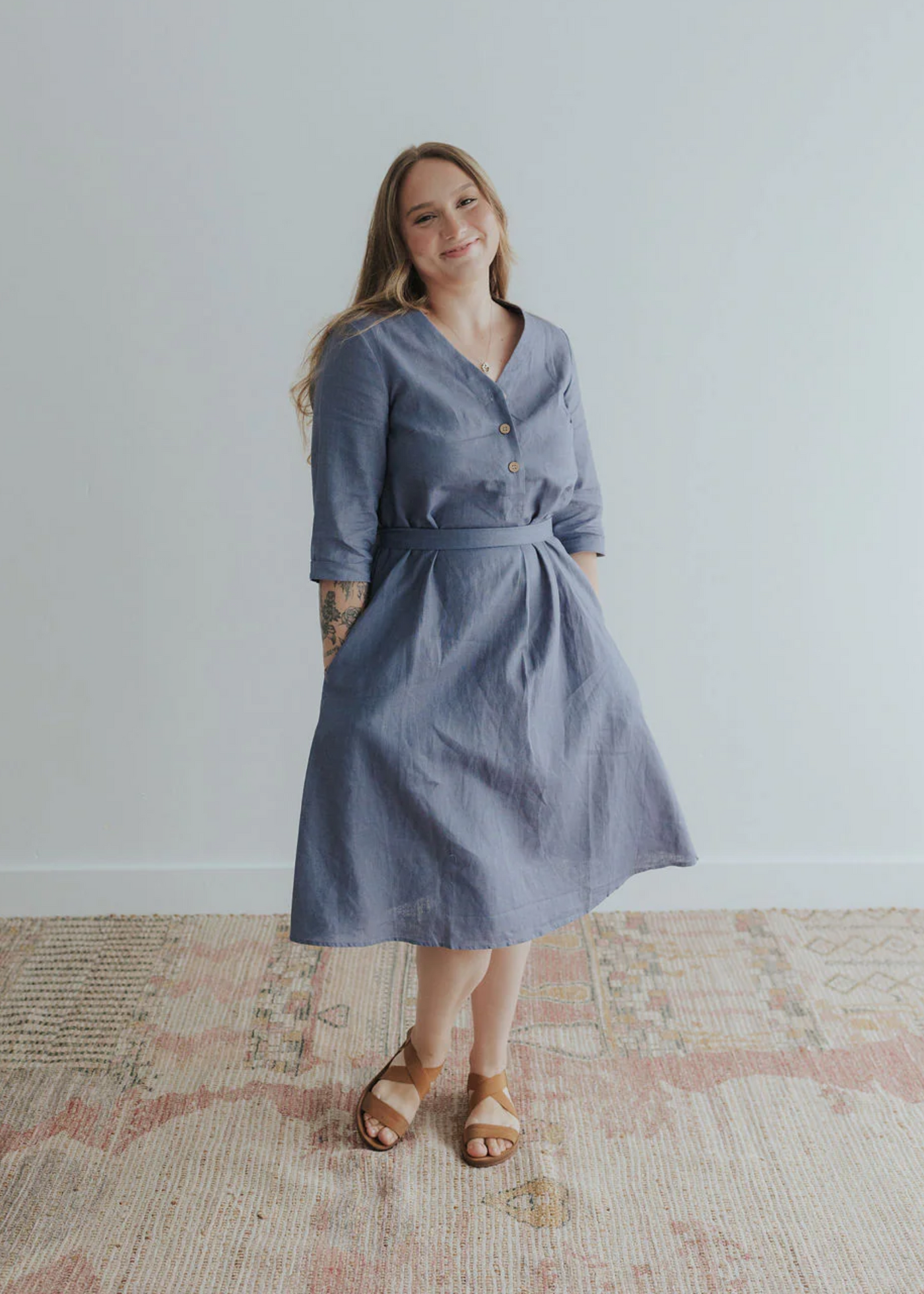 Woman smiling in a blue linen Faye Dress with V-neck buttons, waist tie, and side pockets.