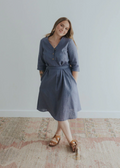 Smiling woman in a blue linen Faye Dress with V-neck buttons, waist tie, and side pockets.
