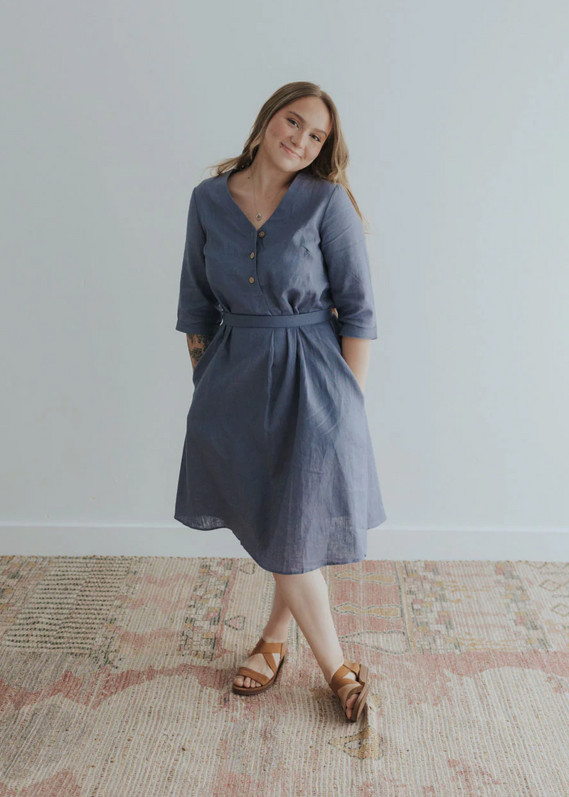 Smiling woman in a blue linen Faye Dress with V-neck buttons, waist tie, and side pockets.