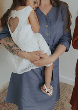 Faye Dress in blue softened linen, V-neck, front buttons, and waist tie, worn by a woman holding a child.