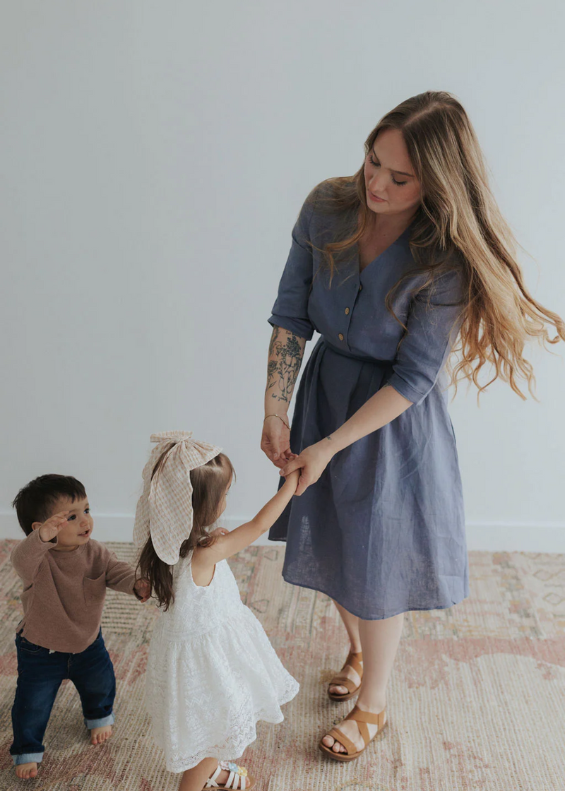 Woman in a blue linen Faye Dress with front buttons and waist tie, holding hands with two small children.