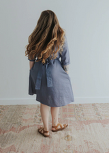 Woman from behind wearing a blue linen Faye dress with a tied waist, side pockets, and brown sandals.
