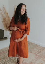 Pregnant woman in a rust linen Faye Dress with front buttons, waist tie, and side pockets.