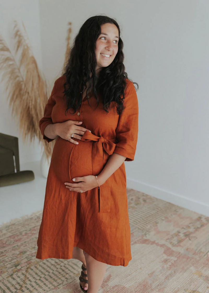 Pregnant woman in a rust linen Faye Dress with front buttons, waist tie, and side pockets.