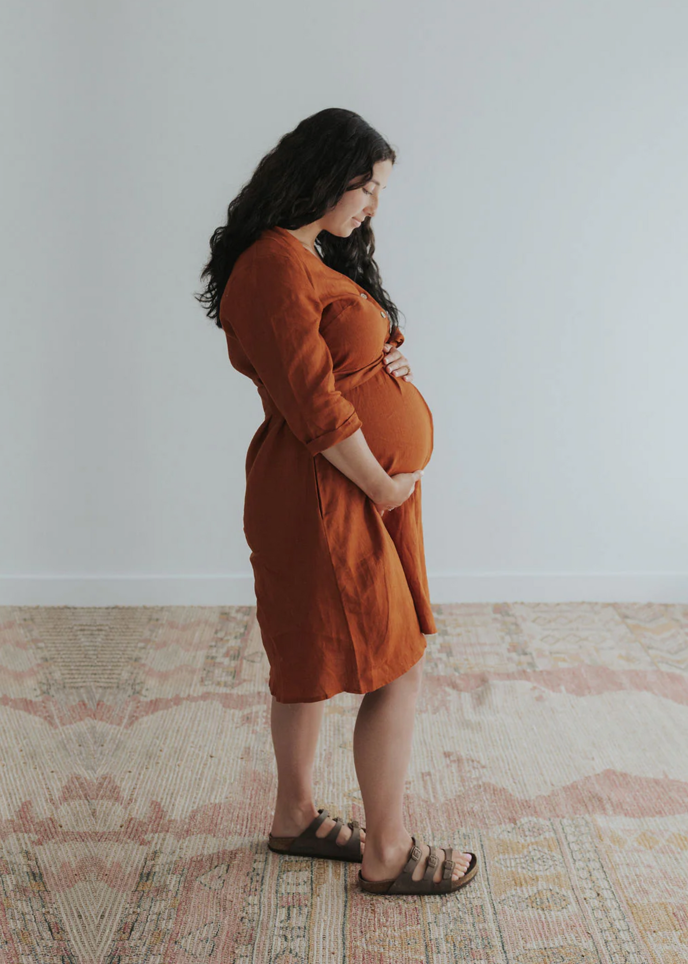 Pregnant woman in a rust linen Faye Dress with front buttons, v-neck, and side pockets.