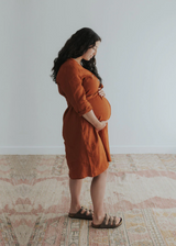 Pregnant woman in a rust linen Faye Dress with front buttons, v-neck, and side pockets.