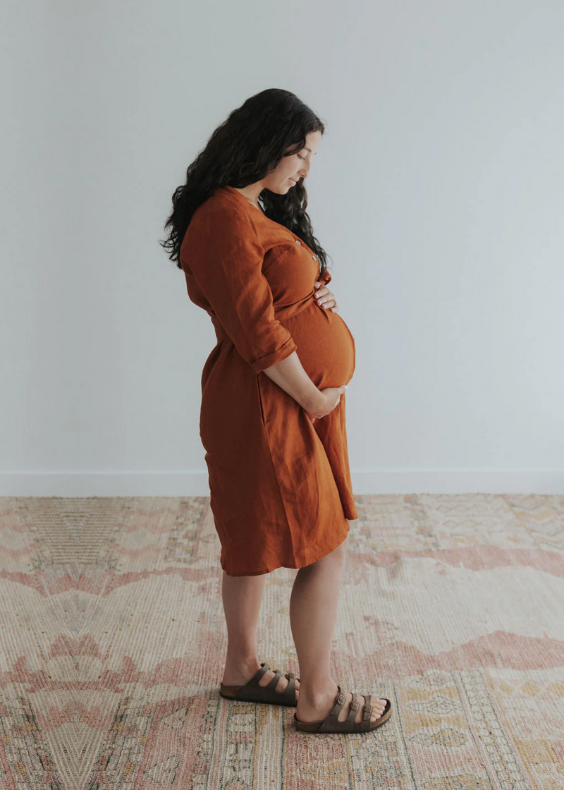 Pregnant woman in a rust linen Faye Dress with front buttons, v-neck, and side pockets.