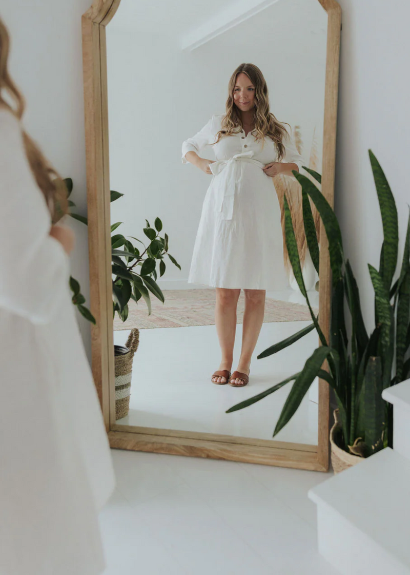 Woman in a white linen Faye Dress with front buttons and waist tie, reflected in a wooden mirror.