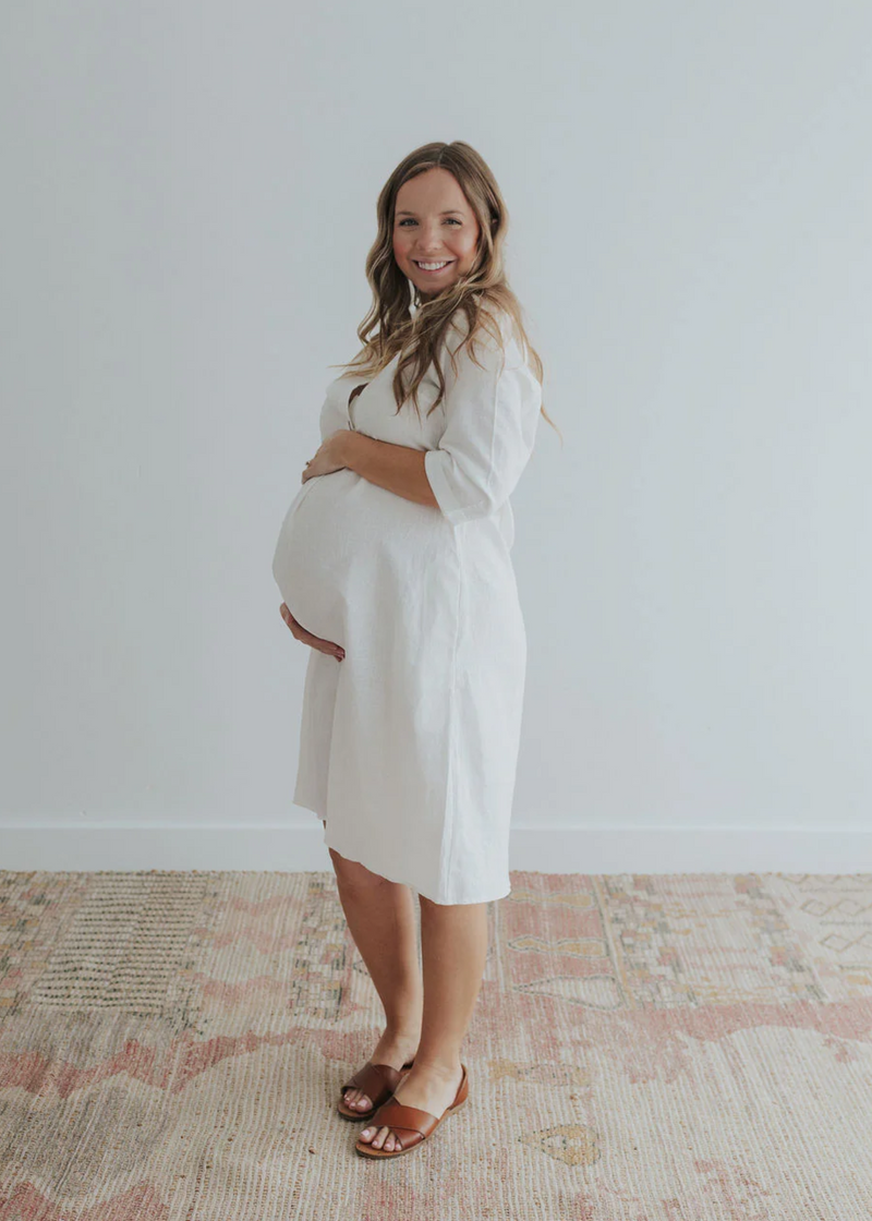 Smiling pregnant woman in a white linen Faye Dress with front buttons, v-neck, and a-line silhouette.