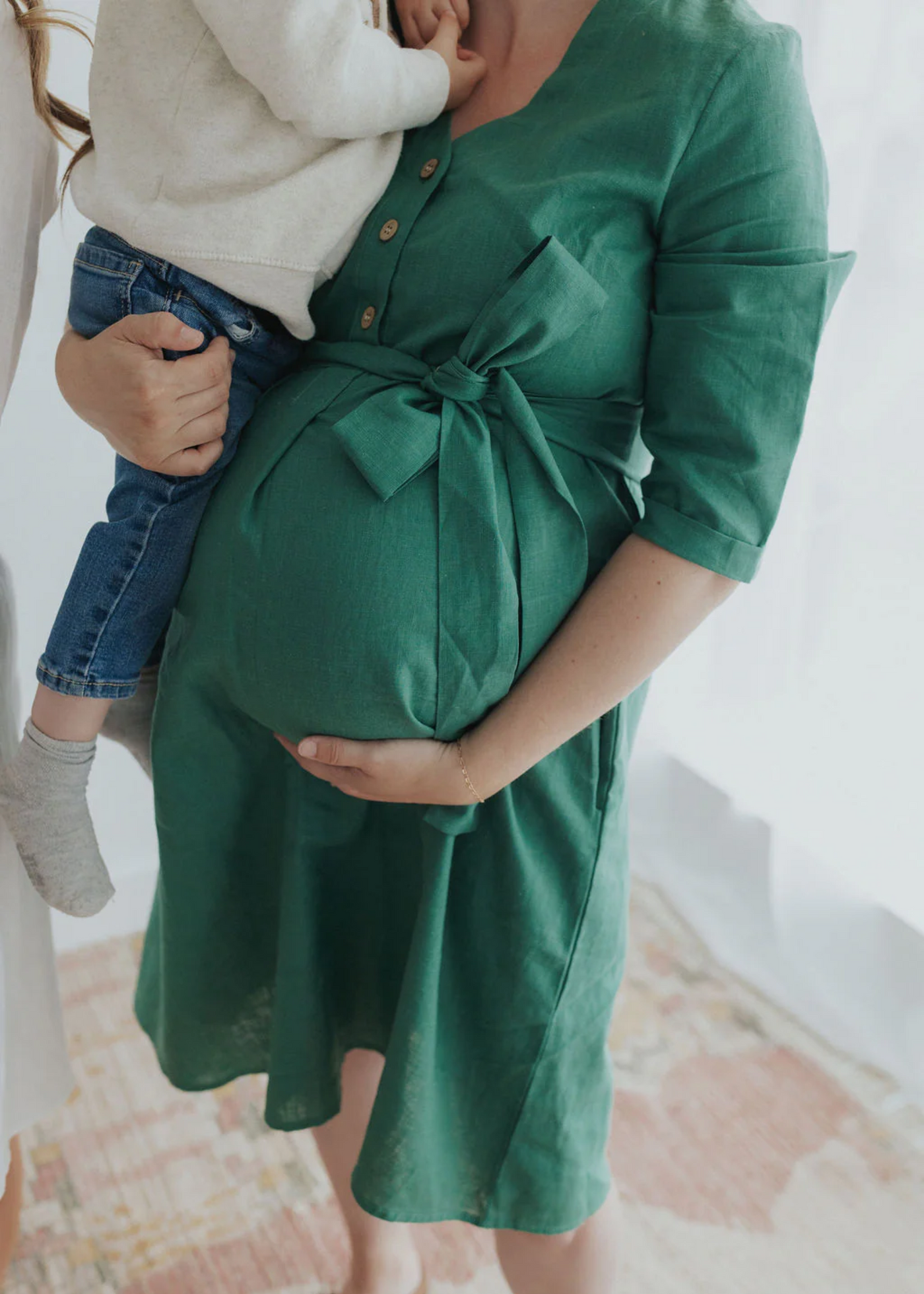 Pregnant woman in a green linen Faye Dress with a tied waist and V-neck button top, holding a child.