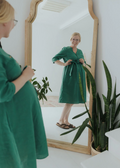 Pregnant woman smiling in a mirror, wearing a green linen Faye Dress with a tied waist and front buttons.
