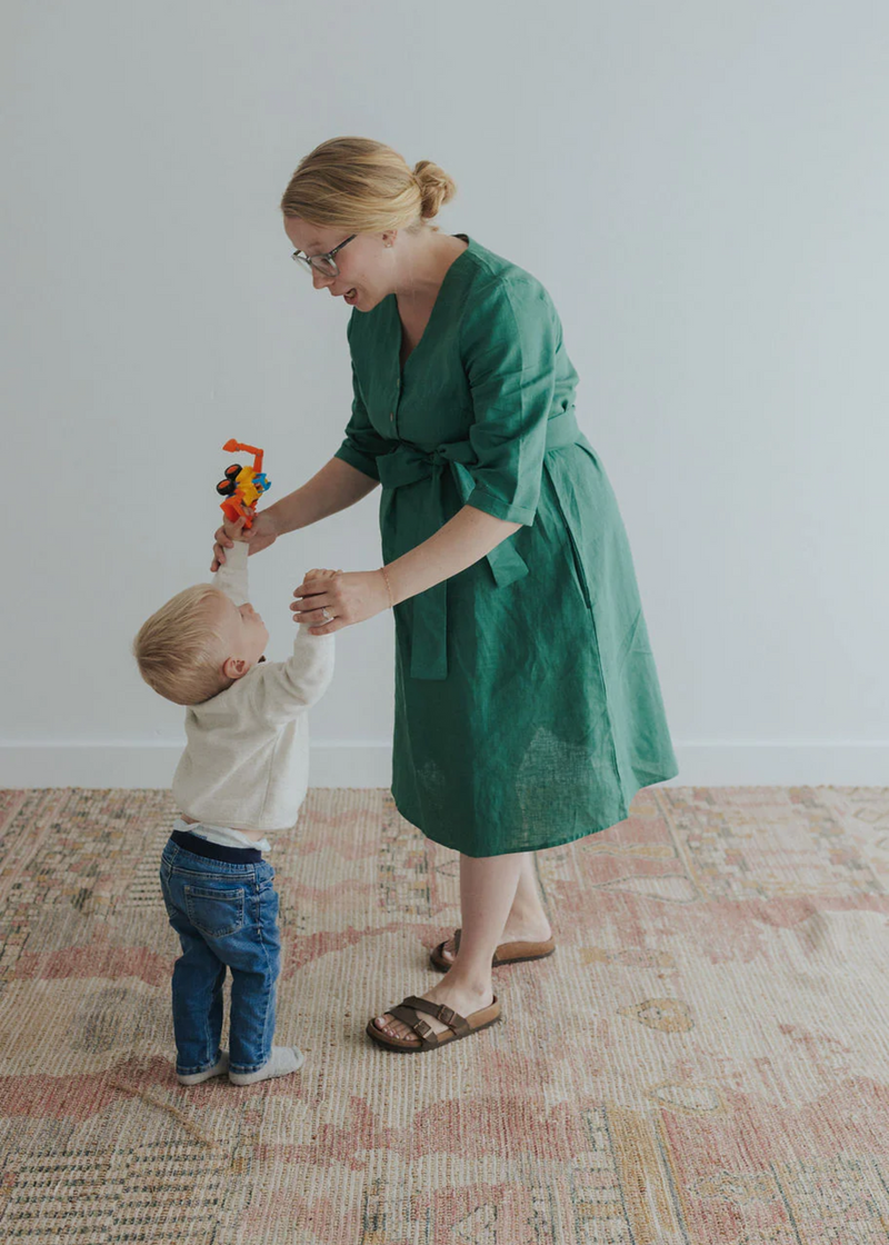 Woman in a green linen Faye Dress with V-neck, front buttons, and waist tie, holding a toddler's hands.