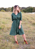 Faye Dress: Woman in a dark green linen dress with front buttons, V-neck, waist tie, and side pockets.