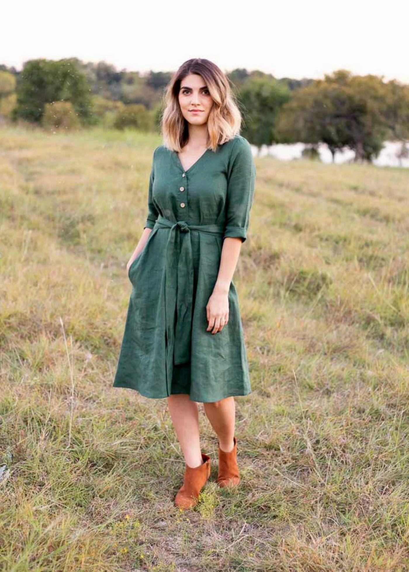 Woman in a dark green linen Faye dress with a V-neck, front buttons, waist tie, and side pockets.