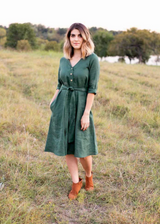 Woman in a dark green linen Faye dress with a V-neck, front buttons, waist tie, and side pockets.