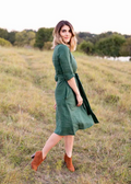 Woman in a dark green linen Faye Dress with a tie waist, V-neck buttons, and side pockets, standing in a field.