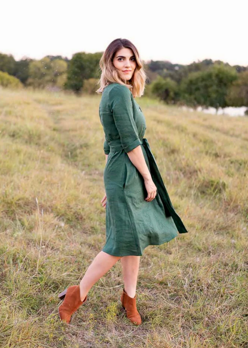 Woman in a dark green linen Faye Dress with a tie waist, V-neck buttons, and side pockets, standing in a field.