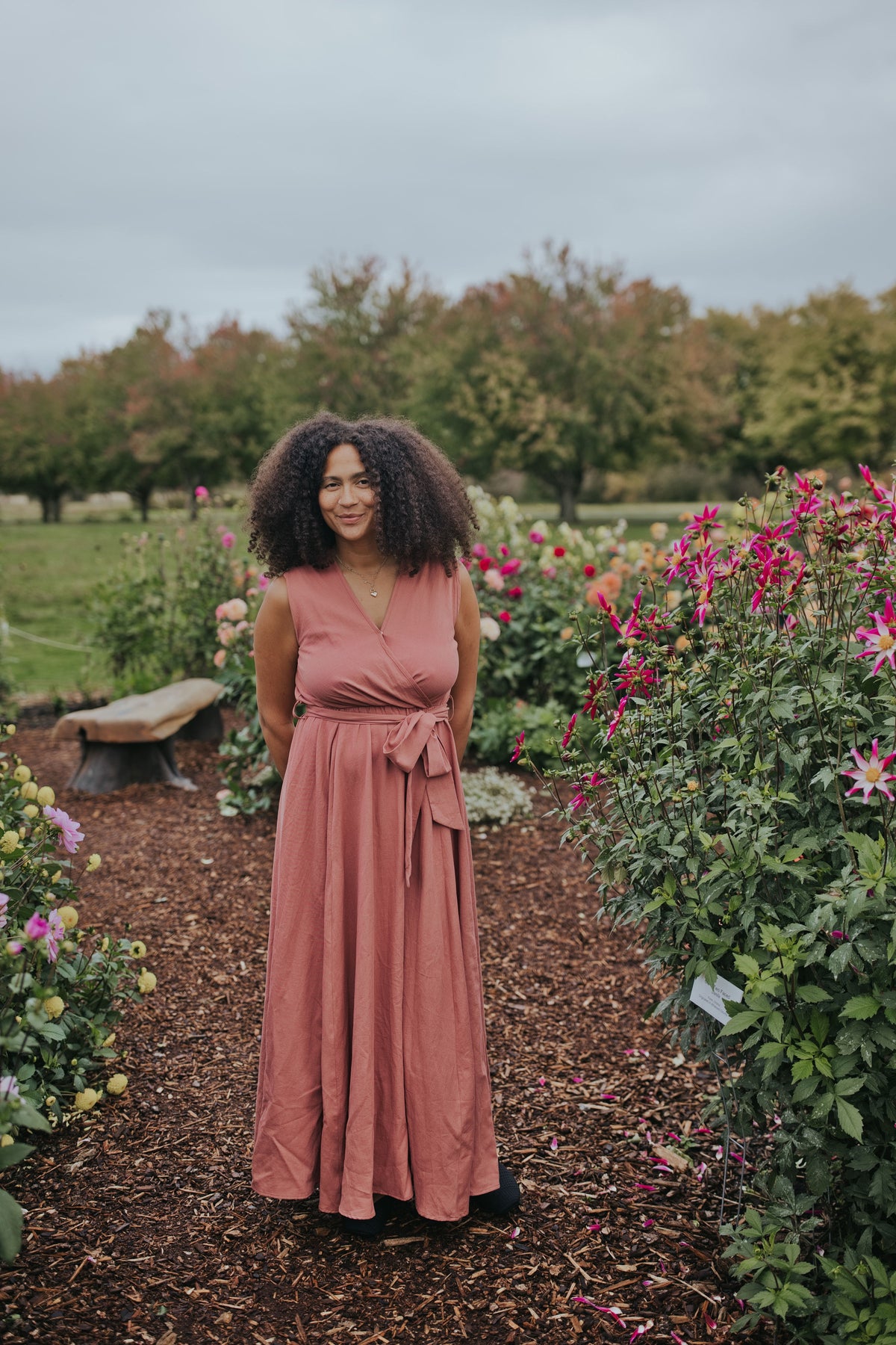 Woman in the Dusty Rose Iris Sleeveless Wrap Dress, front view, full length in a dahlia garden