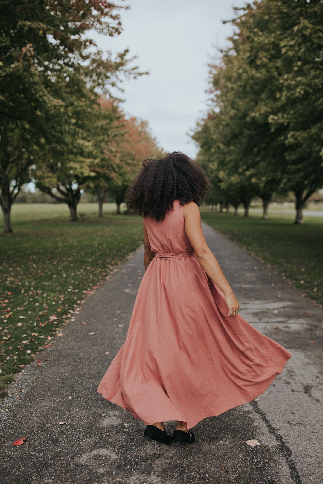 Back view of the Dusty Rose Iris Sleeveless Wrap Dress, skirt in motion on a tree-lined path