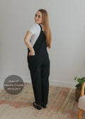Woman in black linen/cotton Journey Overalls with adjustable straps, deep pockets, and side buttons.