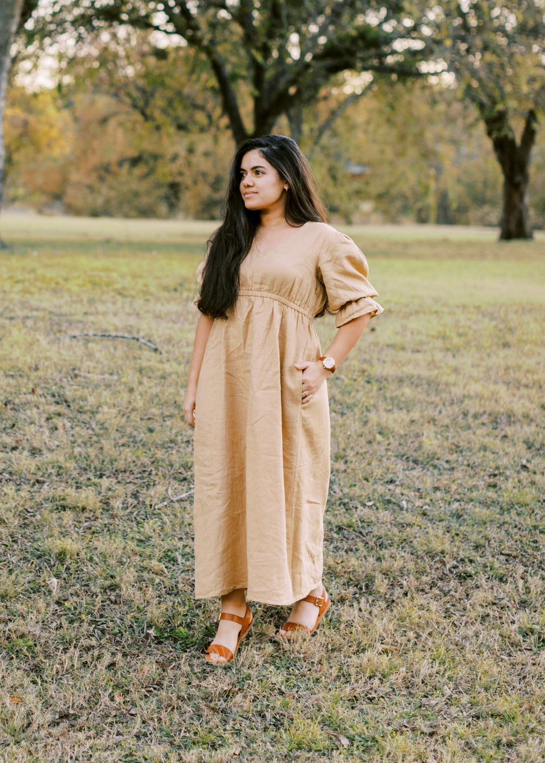 Woman in a long, flowing tan lyocell Julia Dress with puff sleeves and pockets, standing outdoors.