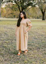 Woman in a long, flowing tan lyocell Julia Dress with puff sleeves and pockets, standing outdoors.