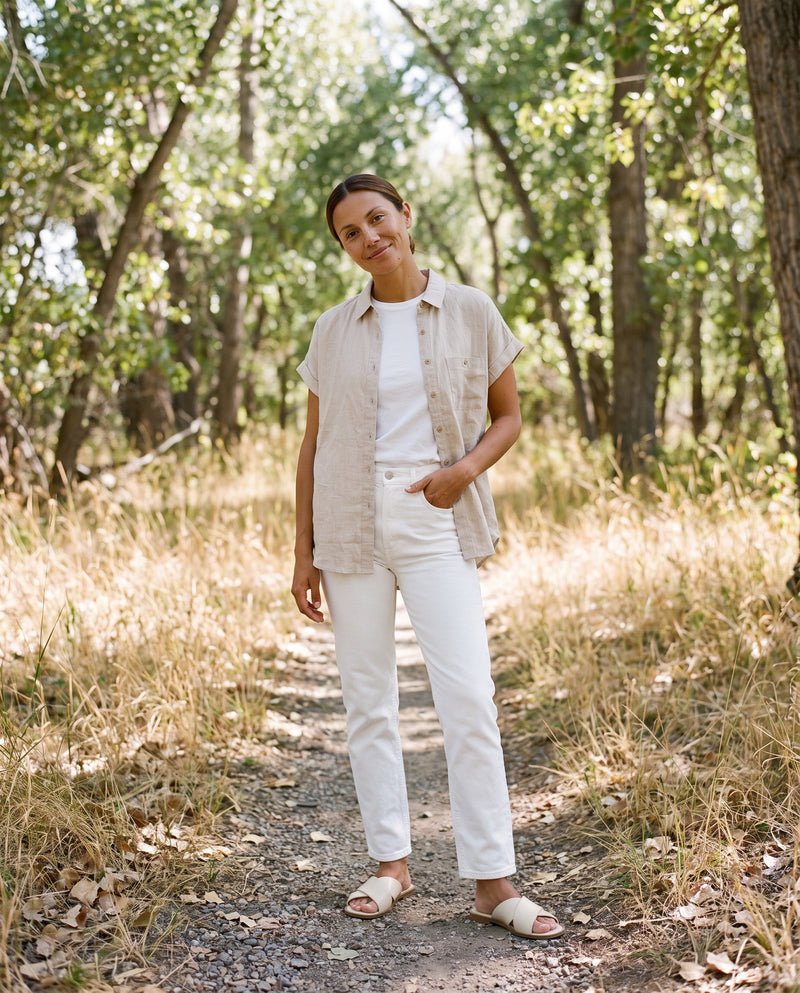 Woman wearing the Nora Camp Shirt in Natural sandwashed linen open over a white tee on a wooded trail