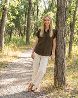 Woman wearing the Nora Camp Shirt in Olive sandwashed linen with cream linen trousers on a wooded trail