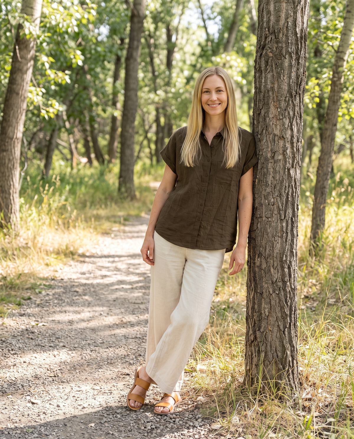 Woman wearing the Nora Camp Shirt in Olive sandwashed linen with cream linen trousers on a wooded trail