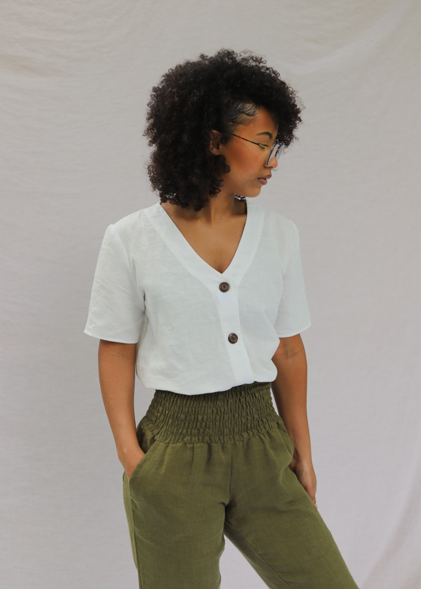 Woman wearing a white linen-cotton Olivia button-down top with a V-neck and brown buttons, paired with olive green pants.