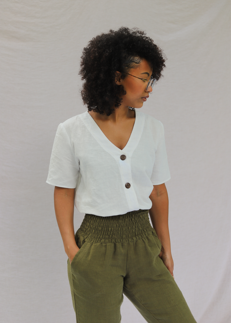 Woman wearing a white linen-cotton Olivia button-down top with a V-neck and brown buttons, paired with olive green pants.