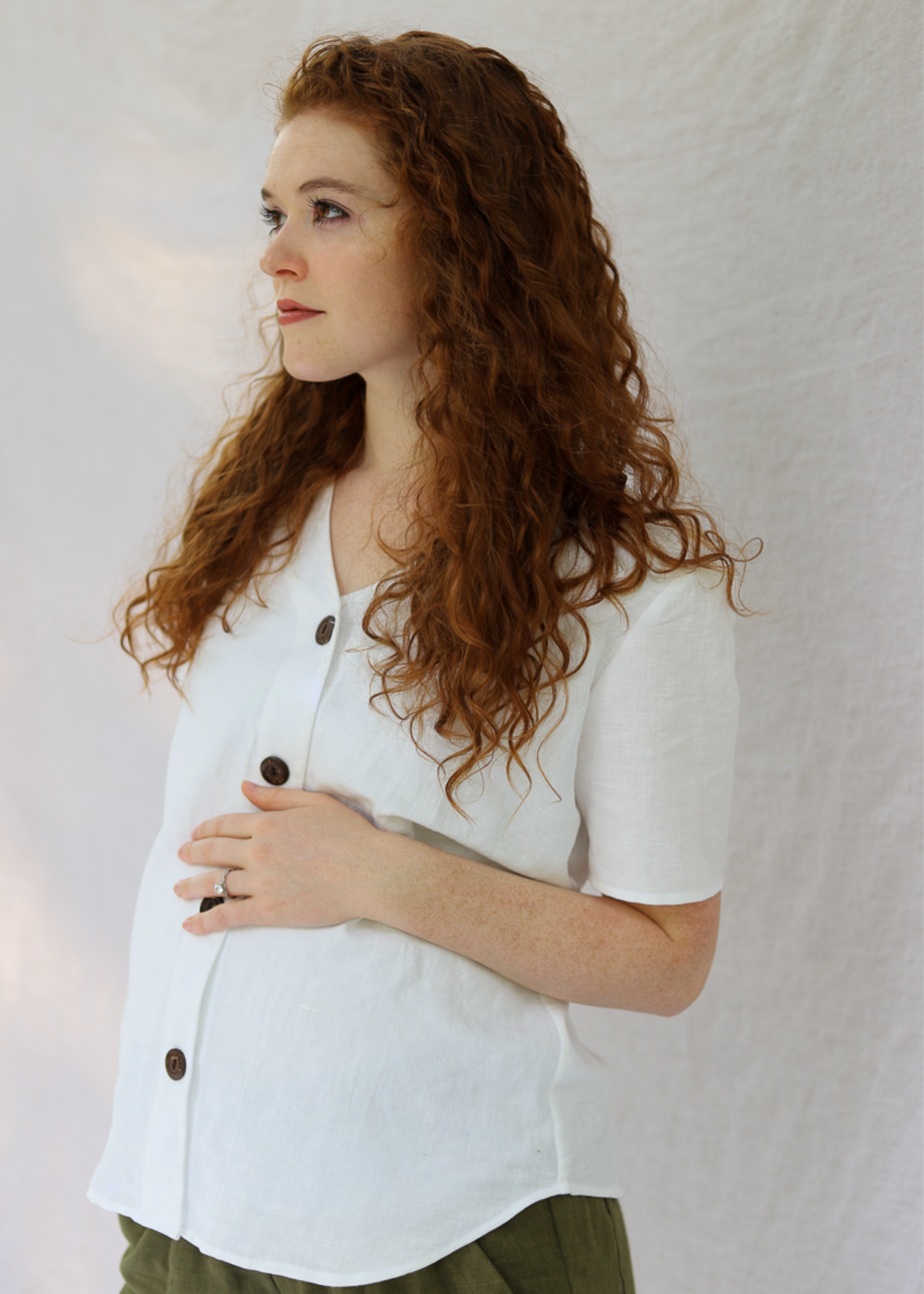 Pregnant woman in a white linen-cotton Olivia button-down top with brown buttons and a V-neck.