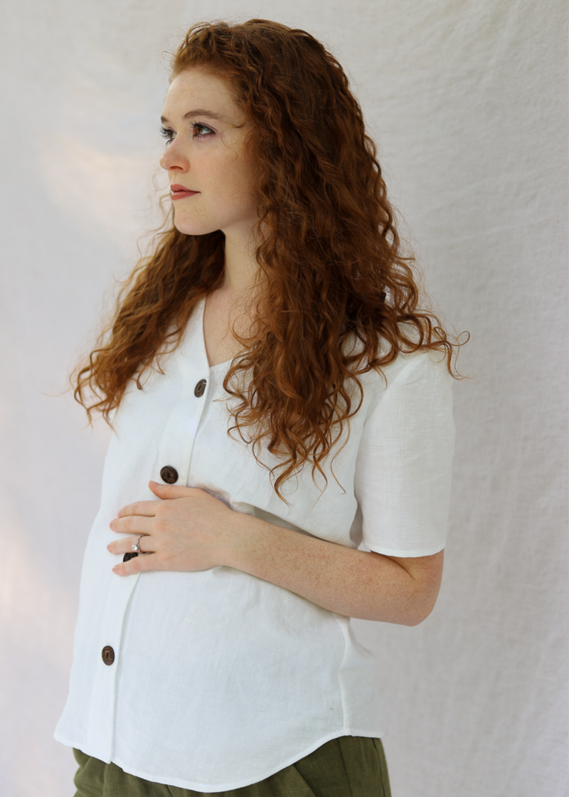 Pregnant woman in a white linen-cotton Olivia button-down top with brown buttons and a V-neck.