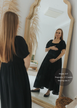 Pregnant woman in a black cotton gauze square neck maxi dress with puffed sleeves, smocked bodice, and tiered skirt.