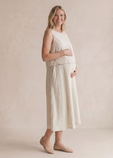 Smiling pregnant woman in a cream linen sleeveless nursing dress with pockets, holding her belly.