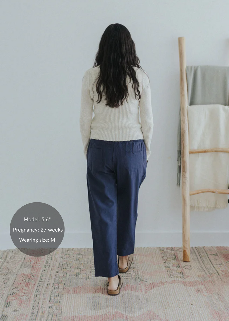 Rear view of a pregnant woman in a cream sweater and navy straight-leg cotton/linen trousers.