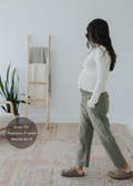 Pregnant woman in olive green straight leg cotton/linen trousers with an elastic waist, paired with a cream cardigan.