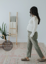 Pregnant woman in olive green straight leg cotton/linen trousers with an elastic waist, paired with a cream cardigan.