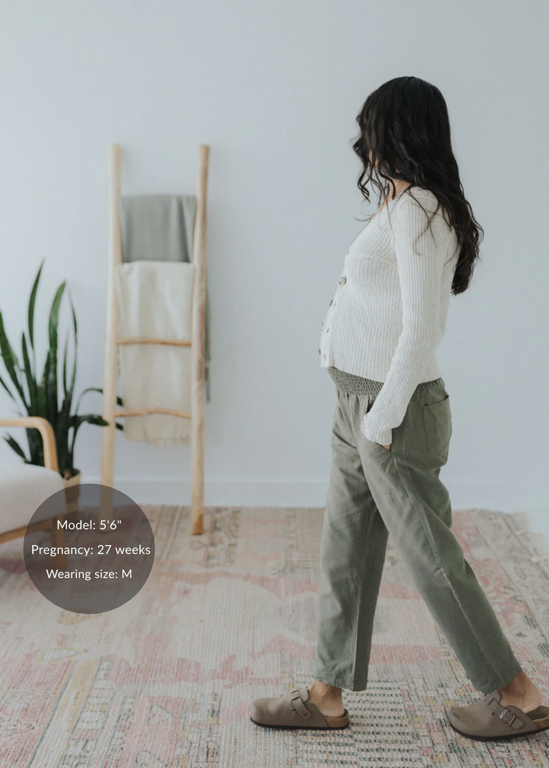 Pregnant woman in olive green straight leg cotton/linen trousers with an elastic waist, paired with a cream cardigan.