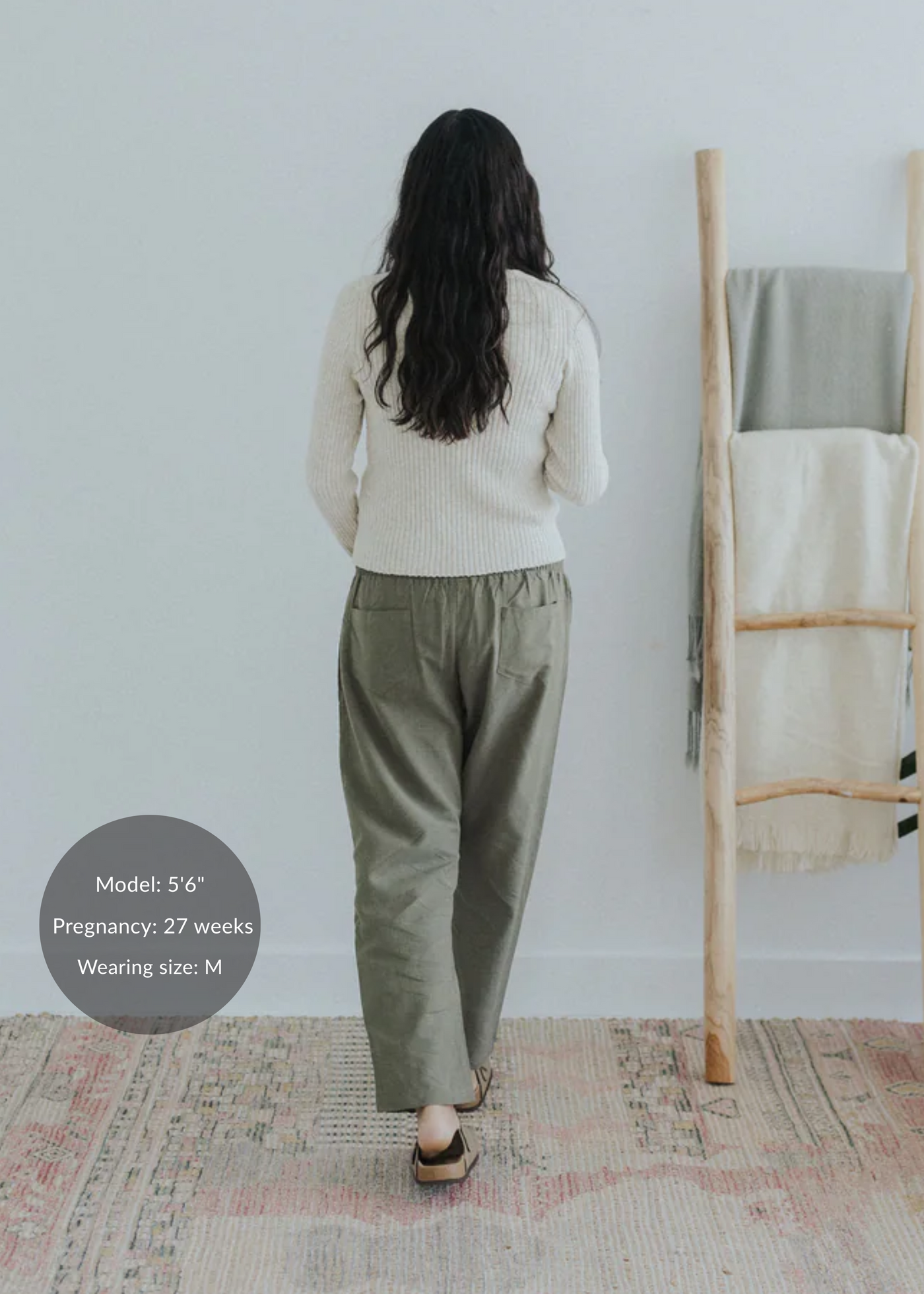 Pregnant model from behind wearing olive straight leg cotton/linen trousers and a cream sweater.