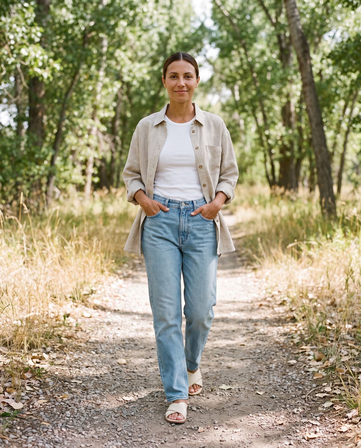 Woman wearing the Sunroom Shirt in Natural sandwashed linen open over a white tank with jeans on a wooded trail