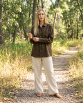 Woman wearing the Sunroom Shirt in Olive sandwashed linen with cream linen trousers on a wooded trail