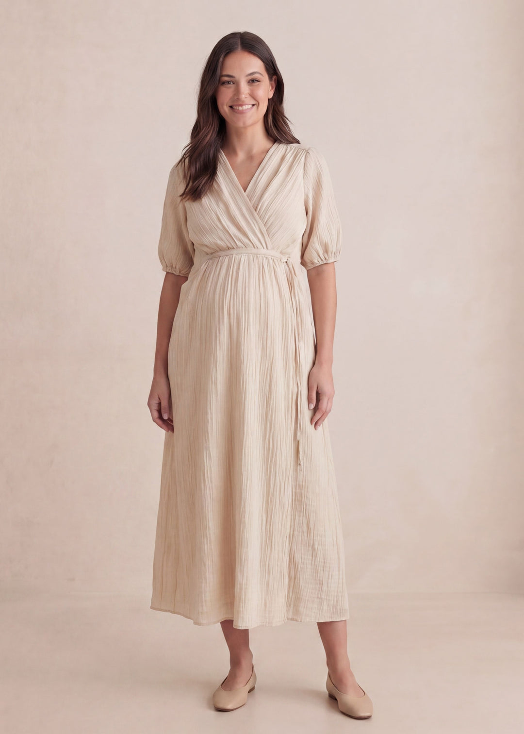 Smiling woman wears a beige textured crossover maternity dress with puff sleeves and a tie waist.
