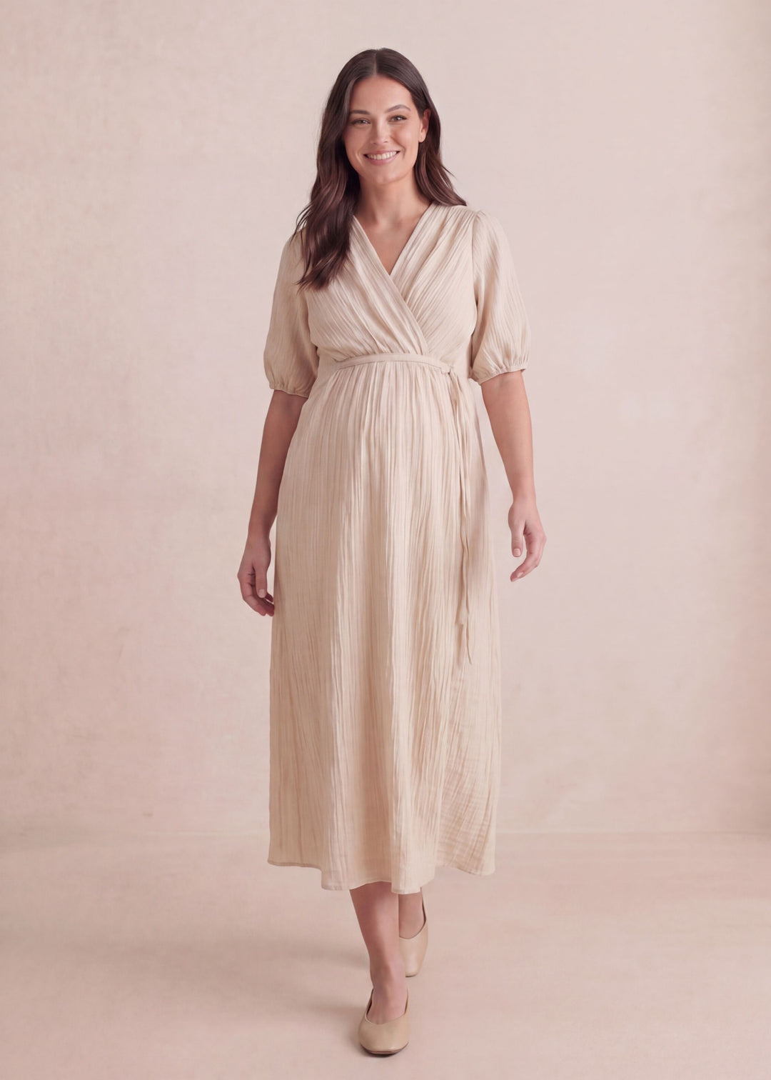 Smiling woman in a textured beige crossover maternity dress with a tie waist and puff sleeves.