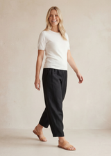 Smiling woman in black linen/cotton barrel pants with deep pockets and a white top, walking right.