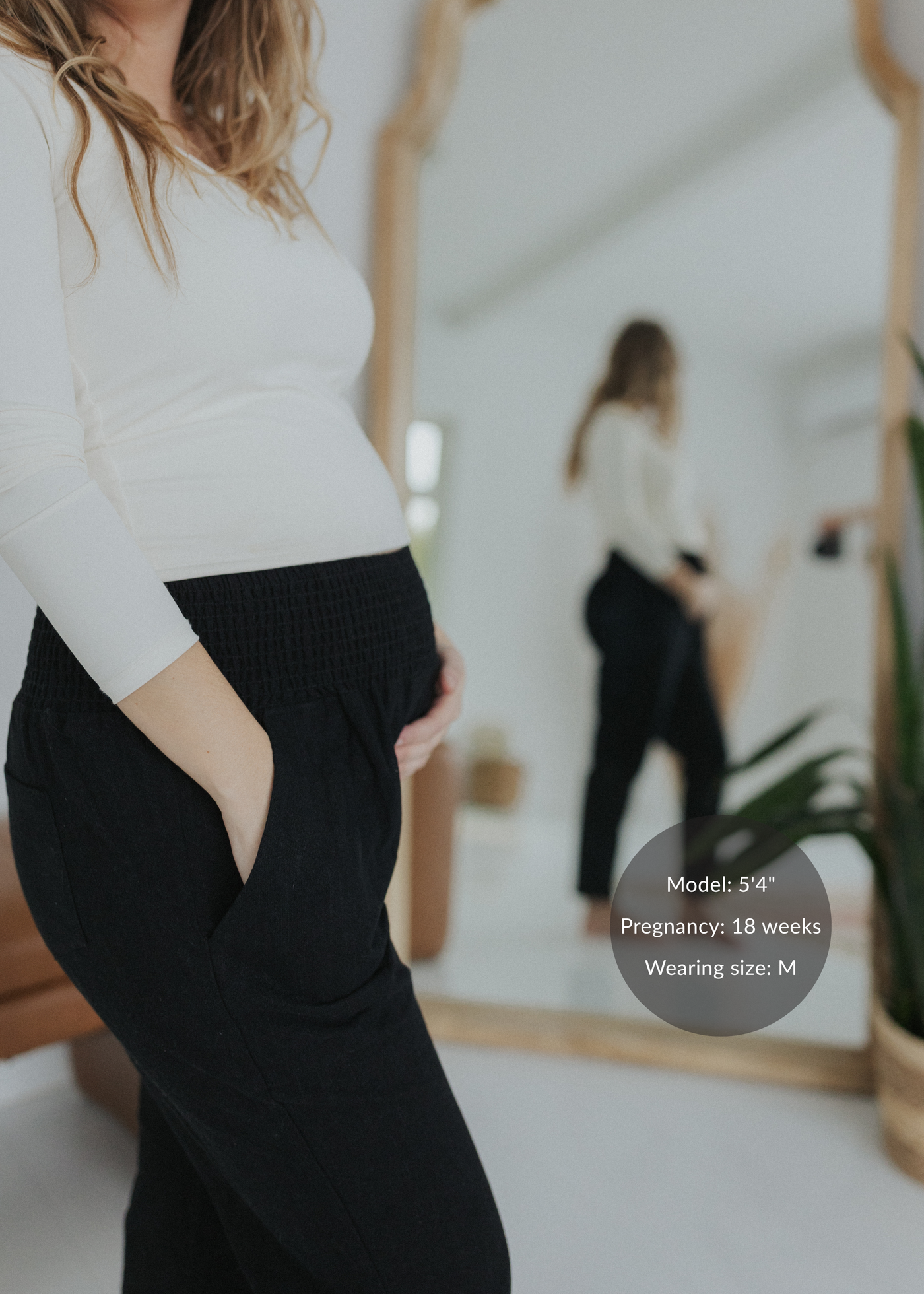 Pregnant woman in black smocked linen pants with pockets and a cream long-sleeve top.